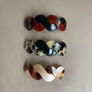 May include: Three hair barrettes with a wavy, twisted design. The top barrette features a glossy black and reddish-brown pattern. The middle barrette has a black and cream spotted print. The bottom barrette displays a smooth cream and brown swirl.