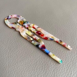 May include: Two colorful hair pins with a marbled pattern. The pins feature a variety of colors, including red, blue, yellow, and white. The pins are made of a glossy material and have a unique shape, with a loop at the top and a forked design.