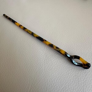 May include: A hair stick with a tortoiseshell pattern. The stick is long and slender, with a rounded tip and a loop at the end. The colors are black, brown, and yellow.