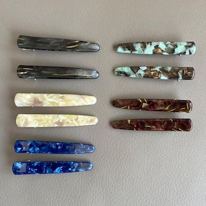 May include: A set of ten rectangular hair clips arranged in two columns. The clips feature various marbled patterns in colors including black, cream, blue, mint green, and dark brown. Each clip has a smooth, glossy finish and a metal hinge mechanism.