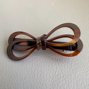 May include: Brown tortoiseshell hair clip in a bow shape. The clip has a barrette style clasp and is made of a glossy, patterned material. The bow design features rounded loops and a central knot.