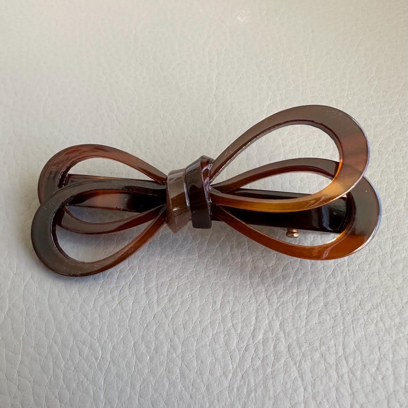 May include: Brown tortoiseshell hair clip in a bow shape. The clip has a barrette style clasp and is made of a glossy, patterned material. The bow design features rounded loops and a central knot.