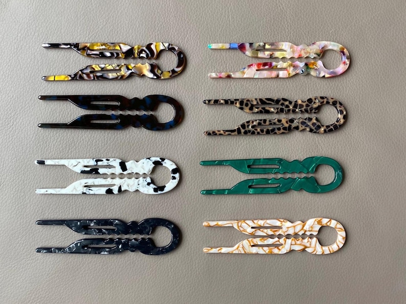 May include: An assortment of eight hair clips in various colors and patterns. The clips feature a unique design with two prongs and a circular handle. Colors include black, white, green, and tortoiseshell patterns. The clips are made of a glossy material.