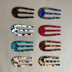 May include: Collection of eight hair forks in various colors and patterns. The forks feature a three-prong design and are made of a glossy, marbled material. Colors include tortoiseshell, blue, white, red, teal, brown, and a multicolored option.