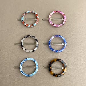 Puede incluir: Seis horquillas de colores con diseños circulares. Cada horquilla presenta un patrón diferente: rojo, azul y blanco; rosa y morado; blanco y negro; azul y blanco; y carey. Las horquillas tienen un cierre plateado tipo pasador.