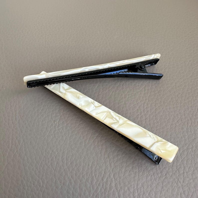 Pu&ograve; includere: Due fermagli per capelli con un design bianco perlaceo e nero. I fermagli hanno una forma rettangolare con una superficie strutturata. La chiusura in metallo &egrave; nera.