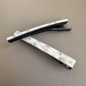 Pu&ograve; includere: Due fermagli per capelli rettangolari. I fermagli hanno una base in metallo nero e un design marmorizzato bianco e lavanda. I fermagli sono appoggiati su una superficie di colore neutro.