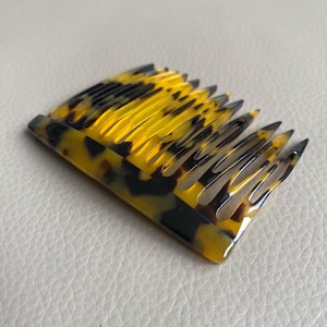 Puede incluir: Un peine para el cabello con un dise&ntilde;o curvo y un patr&oacute;n de carey. El peine presenta tonos amarillos y marrones oscuros alternados, con un acabado brillante. Los dientes est&aacute;n espaciados uniformemente y terminan en punta.