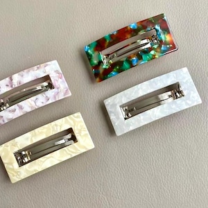 Puede incluir: Cuatro horquillas rectangulares para el cabello en varios colores. Una es una mezcla de rojo, verde y azul, otra es blanca, una tercera es amarillo pálido y la última es una mezcla de rosa y blanco. Cada horquilla tiene un cierre de metal plateado.