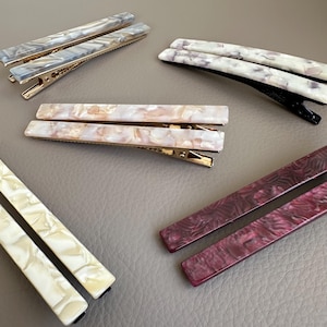 May include: Assortment of rectangular hair clips in various colors, including gray, cream, pink, and burgundy. The clips have a marbled or swirled pattern and gold-colored metal clasps. These accessories are designed to hold hair in place.
