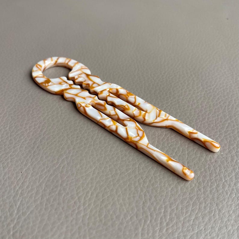 May include: A pair of white and gold hair pins with a marbled pattern. The hair accessories have a looped top and two prongs. The pins are made of a glossy material and are set against a neutral background.
