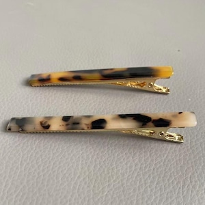 May include: Two rectangular hair clips with a tortoiseshell pattern. One clip has a dark brown and yellow pattern, and the other has a cream and black pattern. Both clips have gold-colored metal clasps.