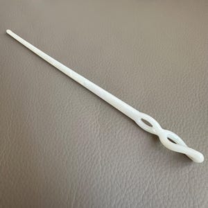 May include: A white hair stick with a twisted design. The hair accessory is made of a solid, smooth material and features a long, tapered shaft. The decorative element has an open, figure-eight design.