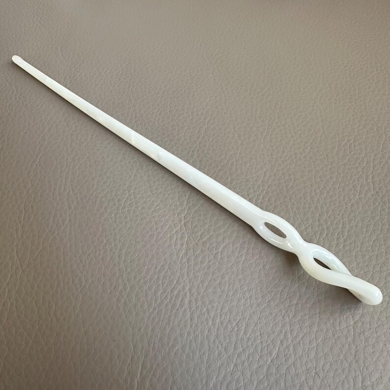 May include: A white hair stick with a twisted design. The hair accessory is made of a solid, smooth material and features a long, tapered shaft. The decorative element has an open, figure-eight design.