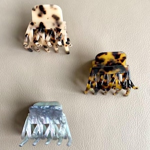 Könnte beinhalten: Drei kleine Haarspangen. Eine hat ein cremefarbenes und braunes Schildpattmuster, eine andere ein gelbes und braunes Schildpattmuster, und die dritte ist perlmuttweiß. Die Spangen sind aus glänzendem, hartem Kunststoff.