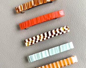 May include: Five rectangular hair barrettes in various colors and patterns. The barrettes include a brown and cream marbled design, a bright orange, a brown and white checkered pattern, a light blue, and a yellow and white striped design.