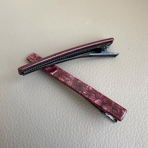 Pu&ograve; includere: Due fermagli per capelli bordeaux con un motivo marmorizzato e chiusure in metallo nero. I fermagli sono rettangolari con una forma leggermente curva. Lo sfondo &egrave; una superficie marrone chiaro neutra.