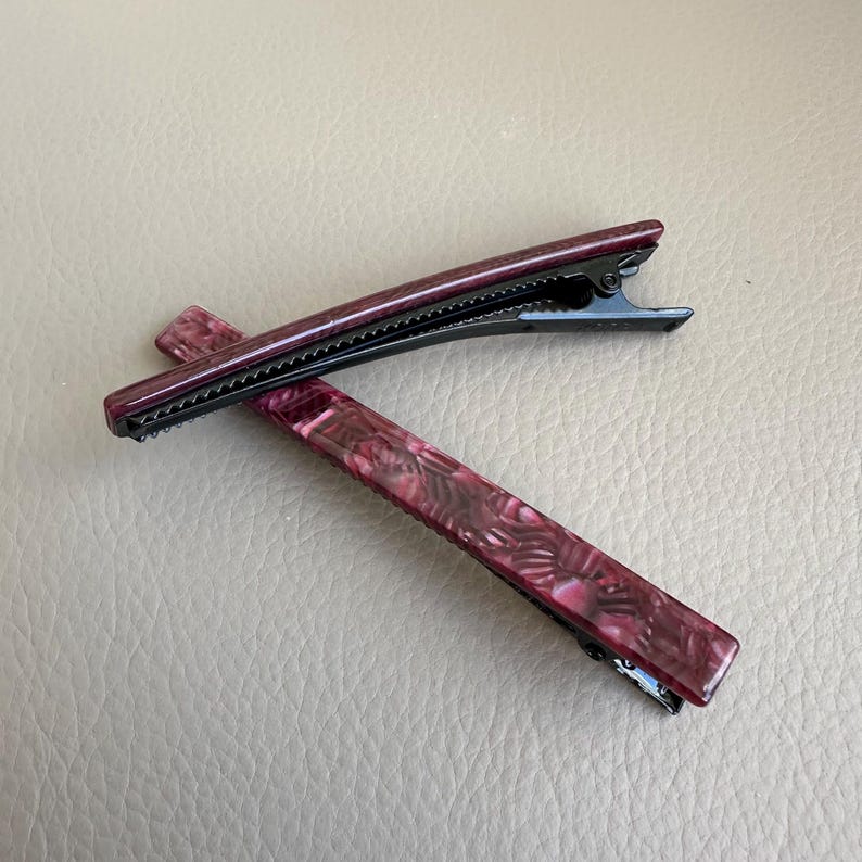 Pu&ograve; includere: Due fermagli per capelli bordeaux con un motivo marmorizzato e chiusure in metallo nero. I fermagli sono rettangolari con una forma leggermente curva. Lo sfondo &egrave; una superficie marrone chiaro neutra.