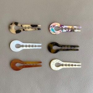 Puede incluir: Seis horquillas decorativas en varios colores y patrones. Los clips son en tonos marrones, blancos y tonos iridiscentes. Cada clip tiene un diseño único, incluyendo un efecto marmolado y un acabado nacarado. Los clips están dispuestos sobre una superficie gris neutra.