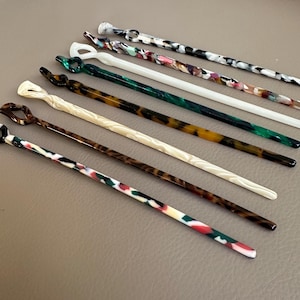 May include: An assortment of colorful hair sticks in various patterns and colors. The hair accessories feature a variety of designs, including marbled, tortoiseshell, and solid colors. The hair sticks are made of a glossy material.