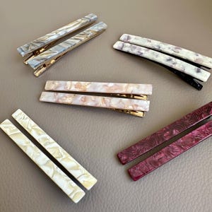 Pu&ograve; includere: Un assortimento di fermagli per capelli rettangolari in vari colori e fantasie. I fermagli presentano chiusure in metallo color oro e sono realizzati con materiali con effetti marmorizzati o a spirale nelle tonalit&agrave; del grigio, bianco, rosa e bordeaux. I fermagli sono disposti su una superficie neutra.