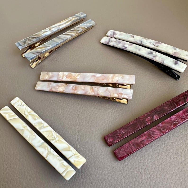 Pu&ograve; includere: Un assortimento di fermagli per capelli rettangolari in vari colori e fantasie. I fermagli presentano chiusure in metallo color oro e sono realizzati con materiali con effetti marmorizzati o a spirale nelle tonalit&agrave; del grigio, bianco, rosa e bordeaux. I fermagli sono disposti su una superficie neutra.