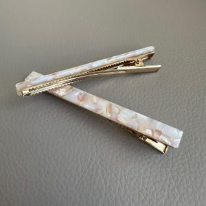 Pu&ograve; includere: Due fermagli per capelli rettangolari con un design marmorizzato perlato nei toni del bianco, crema e pesca. I fermagli hanno una chiusura in metallo color oro. Gli accessori per capelli sono su una superficie grigia.