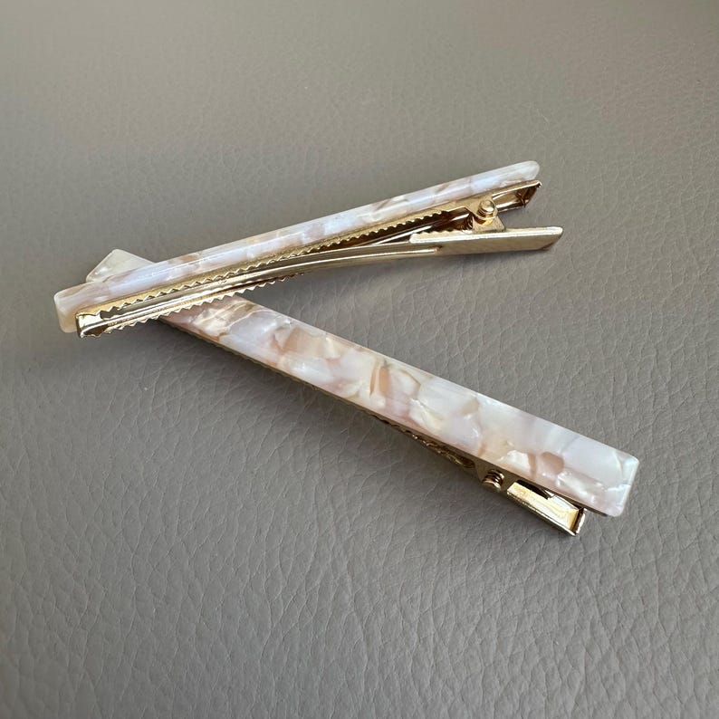 Pu&ograve; includere: Due fermagli per capelli rettangolari con un design marmorizzato perlato nei toni del bianco, crema e pesca. I fermagli hanno una chiusura in metallo color oro. Gli accessori per capelli sono su una superficie grigia.