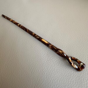 May include: A decorative hair stick with a swirling pattern of brown and tan. The stick has a slender, tapered design with a looped end. The hair accessory is made of a glossy material.