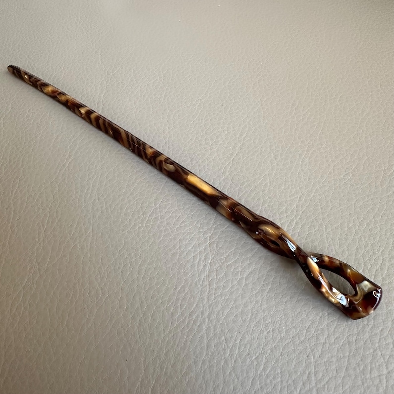 May include: A decorative hair stick with a swirling pattern of brown and tan. The stick has a slender, tapered design with a looped end. The hair accessory is made of a glossy material.