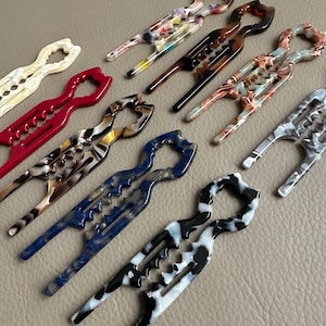 Könnte beinhalten: Eine Sammlung farbenfroher Haarspangen in verschiedenen Mustern und Farben, darunter Rot, Blau und Schildpatt. Die Spangen haben ein einzigartiges, offenes Design mit einem gebogenen Griff und einer Reihe von Zähnen zum Befestigen der Haare.