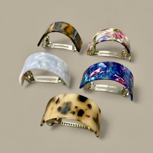 May include: Five colorful hair clips with a curved design. The clips feature various patterns, including tortoiseshell, floral, and marbled effects in shades of blue, pink, and white. Each clip has a silver metal clasp.
