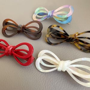 Peut inclure: Cinq pinces à cheveux en forme de nœud papillon de différentes couleurs. Les pinces comprennent un rouge, un écaille de tortue, un marron, un blanc nacré et un clip aux couleurs de l'arc-en-ciel pastel. Chaque pince a un fermoir en métal à l'arrière.