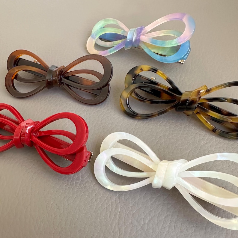 May include: Five bow-shaped hair clips in various colors. The clips include a red, a tortoiseshell, a brown, a pearlescent white, and a pastel rainbow-colored clip. Each clip has a metal clasp on the back.