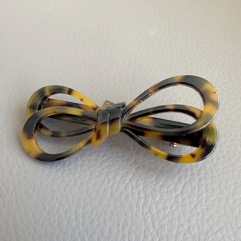 May include: A tortoiseshell-patterned hair clip in a bow shape. The clip features a mix of brown, black, and yellow hues. The bow design is detailed and has a barrette style clasp on the back. This accessory is suitable for various hairstyles.