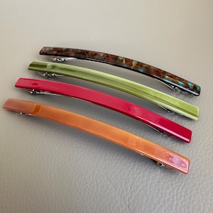 May include: Four colourful hair barrettes in shades of brown, green, red, and orange. Each barrette has a curved rectangular shape with a silver clasp. The barrettes are made of a glossy material.
