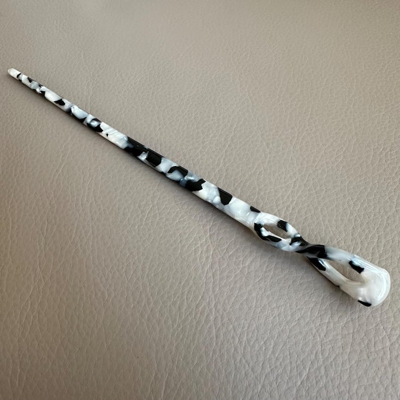 May include: A decorative hair stick with a swirling design. The stick is white with black and gray spots, and has a rounded end. The hair accessory is approximately 7 inches long.
