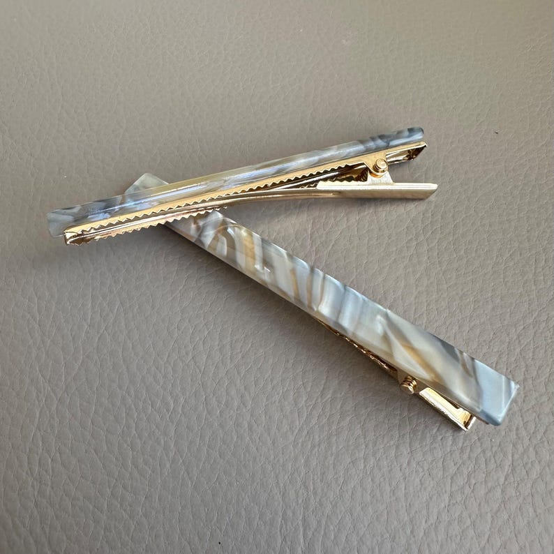 Pu&ograve; includere: Due fermagli per capelli rettangolari con un motivo marmorizzato nei toni del grigio, bianco e beige. I fermagli hanno una chiusura in metallo color oro. I fermagli sono appoggiati su una superficie grigia.