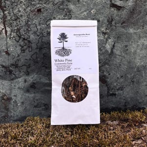 May include: A white paper bag with a brown, dried root inside. The bag has a label that says "Ashwagandha Root" and "White Pine Community Farm" with contact information.