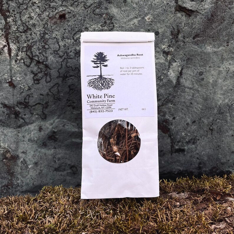 May include: A white paper bag with a brown, dried root inside. The bag has a label that says "Ashwagandha Root" and "White Pine Community Farm" with contact information.