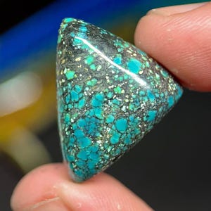 May include: A triangular turquoise gemstone with a speckled pattern of teal, black, and white. The polished stone is held between two fingers, showcasing its vibrant colors and unique texture. The gemstone has a smooth, glossy surface.