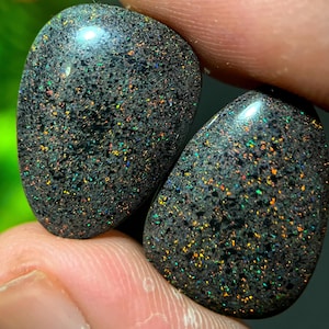 Könnte beinhalten: Zwei polierte schwarze Opal-Edelsteine mit gesprenkeltem Aussehen und schillernden Farbblitzen. Die Opale haben eine Tropfenform und ein glänzendes Finish, das eine Reihe von Farben wie Grün, Orange und Gelb zeigt.