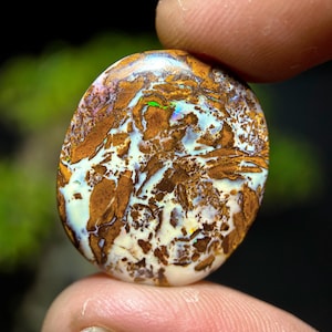 May include: An oval-shaped boulder opal gemstone featuring a polished surface with a complex matrix of dark brown ironstone veins interspersed with iridescent flashes of bright green, blue, and yellow light across the creamy white opal material.