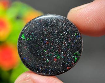May include: A round, black opal gemstone with a speckled appearance. The opal displays flashes of green, red, and blue throughout its surface. The gemstone is held between fingers, with a blurred green and yellow background.