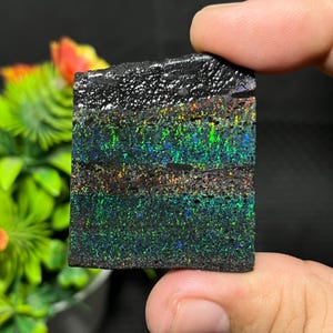 Op de afbeelding: Een rechthoekige zwarte opaal edelsteen met iriserende lagen groen, blauw en oranje. Het gepolijste oppervlak reflecteert licht en toont een spectrum aan kleuren. De opaal wordt vastgehouden, wat de natuurlijke schoonheid en het unieke patroon benadrukt.
