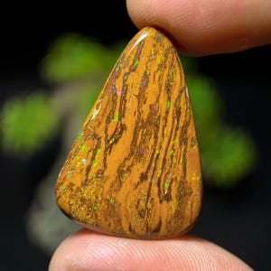 May include: A triangular-shaped boulder opal gemstone featuring a warm, earthy brown matrix with vertical, dark brown veining. The surface displays vibrant, multi-colored flashes of green, yellow, and orange fire throughout the stone's texture.