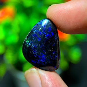 Könnte beinhalten: Ein polierter schwarzer Opal-Edelstein mit leuchtenden blauen und grünen Blitzen. Der Stein wird zwischen zwei Fingern gehalten, wodurch seine glatte, abgerundete Form und das Farbspiel im Inneren zur Geltung kommen.