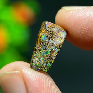 May include: A rectangular, rough-cut opal gemstone with a dark brown matrix. The opal displays vibrant flashes of green, blue, and gold, with a speckled appearance. The gemstone is held between two fingers, showcasing its natural beauty.