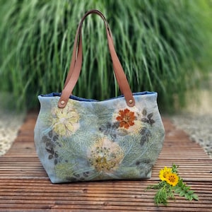 Peut inclure: Un sac cabas en jean bleu clair orné d'un imprimé botanique de fleurs et de feuillages pressés dans des tons jaune, orange et vert foncé. Le sac possède deux anses d'épaule en cuir beige fixées par des rivets métalliques dorés.