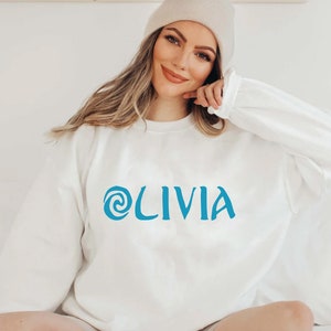 May include: A white sweatshirt with a blue swirl design and the name 'OLIVIA' printed on the front.